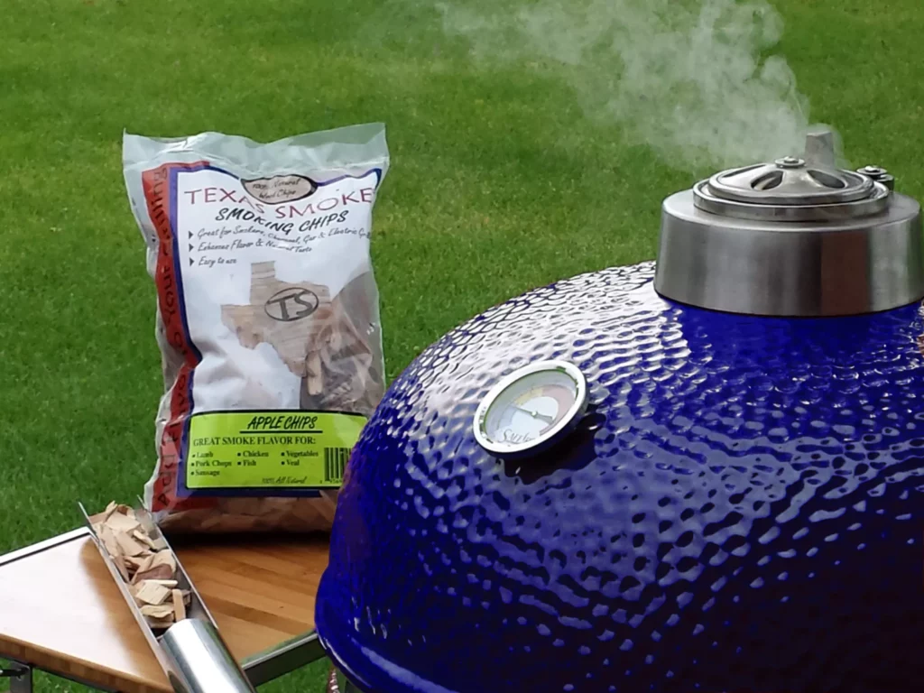 A blue Saffire kamado with steam coming out the top and a bag of wood chips and the smokin' chip feeder tool on the grill's side shelf