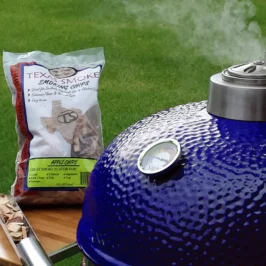 A blue Saffire kamado with steam coming out the top and a bag of wood chips and the smokin' chip feeder tool on the grill's side shelf