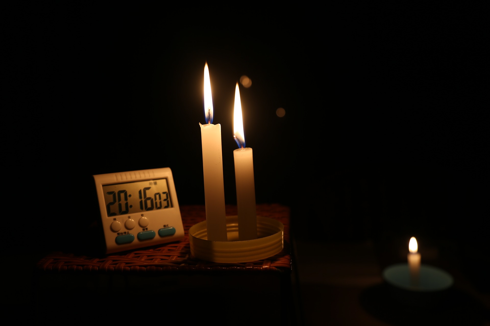 Two emergency candles are lit together on a wicker basket in a pitch-black room. A timer sits on the wicker basket near the candles, with the time set to 20 minutes and 16 seconds.