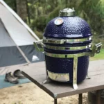 The 13 inch small Saffire ceramic grill sits on a camping table. In the background is a tent and boots sitting on a mat outside the tent.