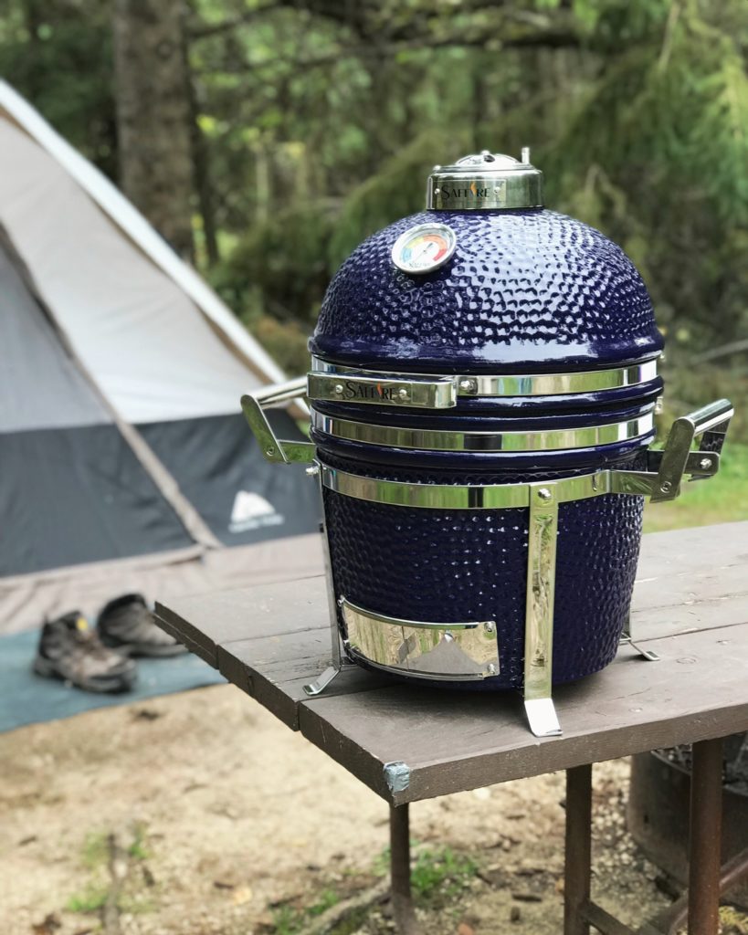 The 13 inch small Saffire ceramic grill sits on a camping table. In the background is a tent and boots sitting on a mat outside the tent.