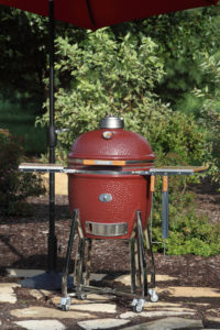 An SG18 Jasper Red Saffire grill sits in a beautiful backyard with an umbrella over it; on one of its side shelves hangs two grilling tools