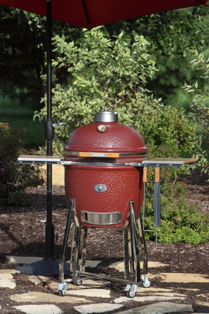 An SG18 Jasper Red Saffire grill sits in a beautiful backyard with an umbrella over it; on one of its side shelves hangs two grilling tools