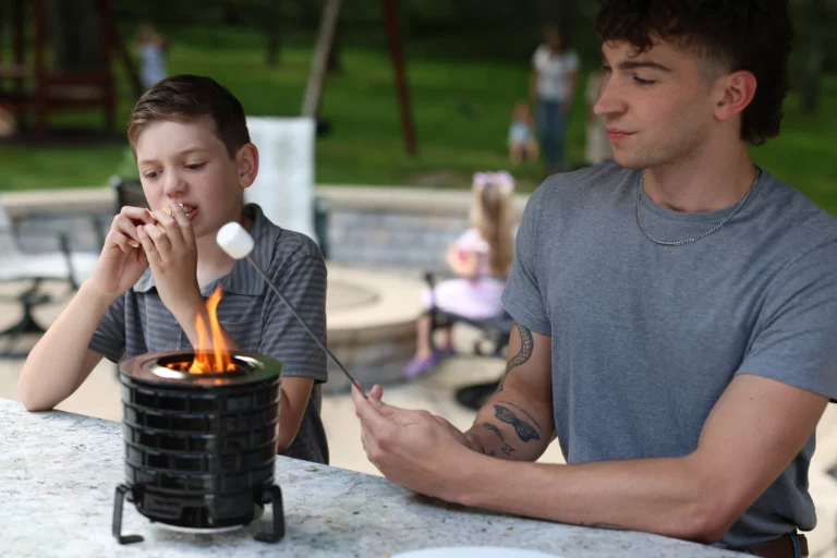 two boys sitting at a table with marshmallows over a fire