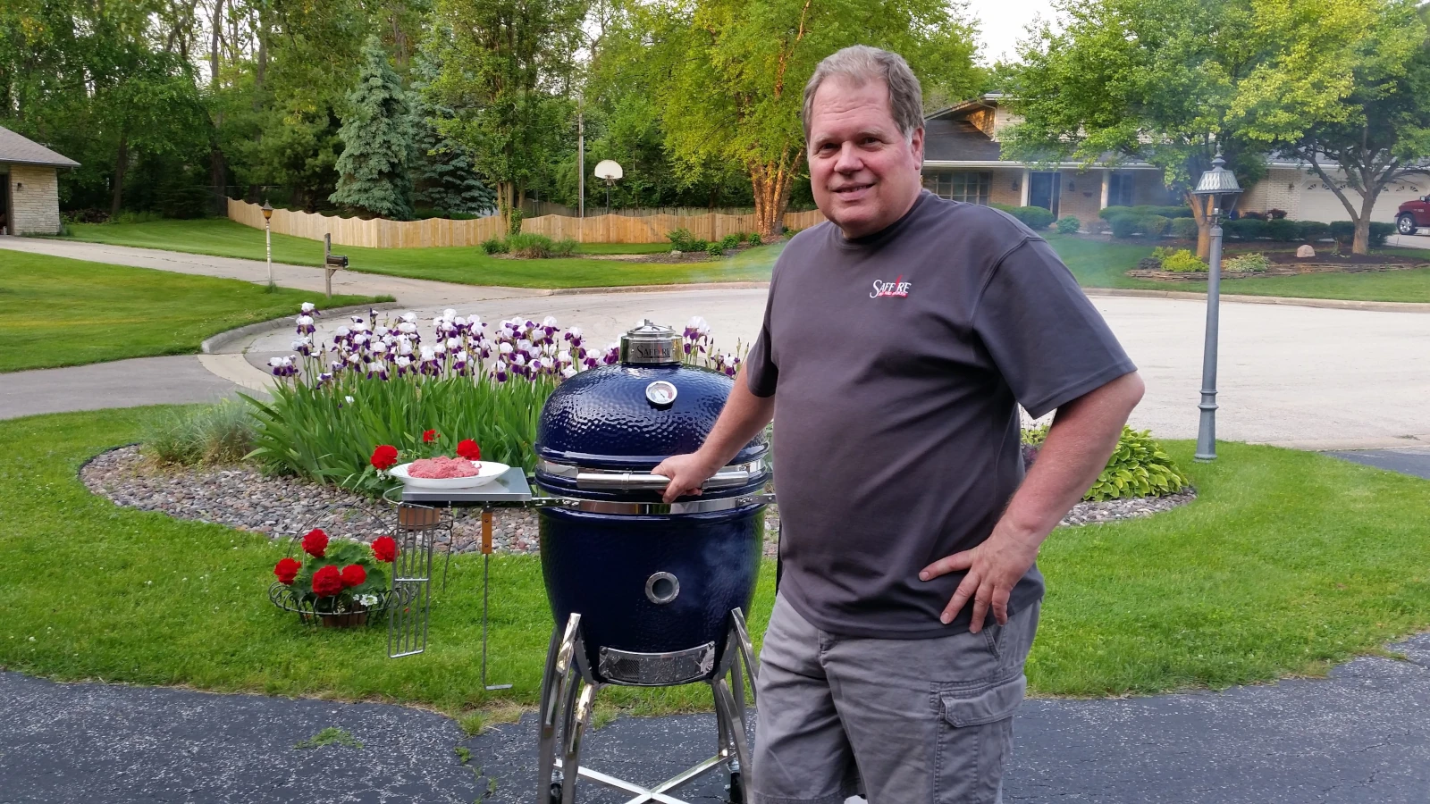 Steve standing in front of his 19" Platinum Saffire Kamado. He has his hand on his hip and the other hand on the grill's handle. There is some ground beef on the grill's side shelf, and smoke is coming out of the open Smokin' Chip Feeder port.